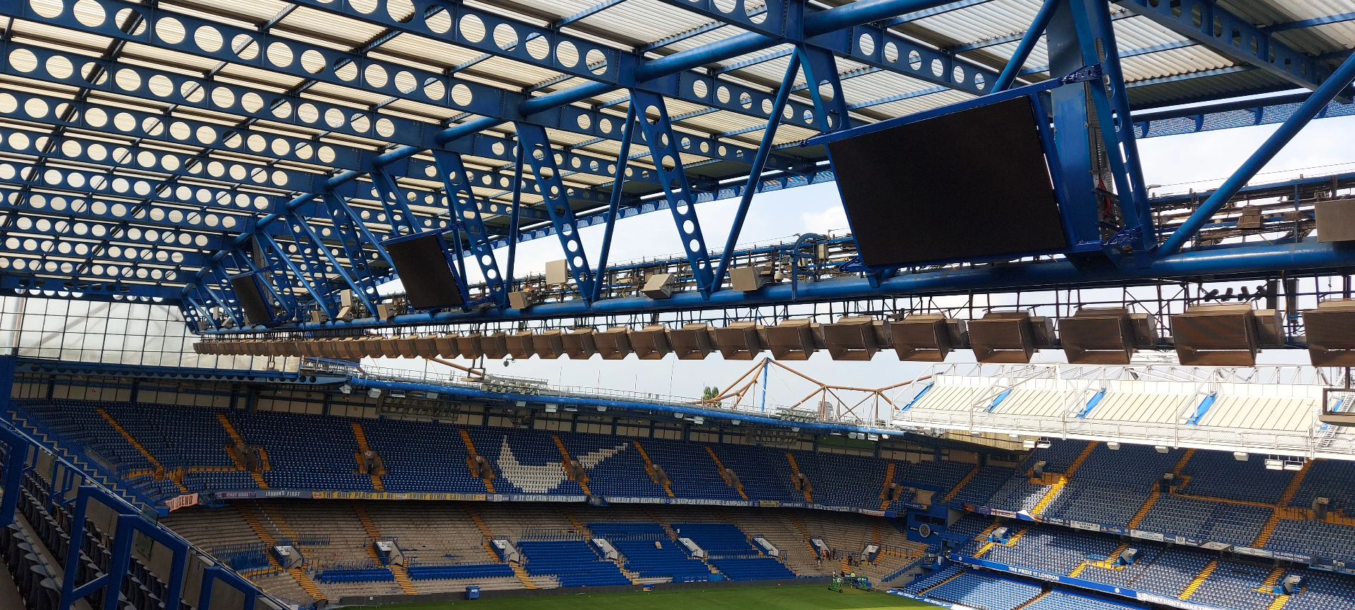 Chelsea FC — under-roof view of Stamford Bridge primary truss with rigged scoreboard screen and PA / lighting array suspended at high level