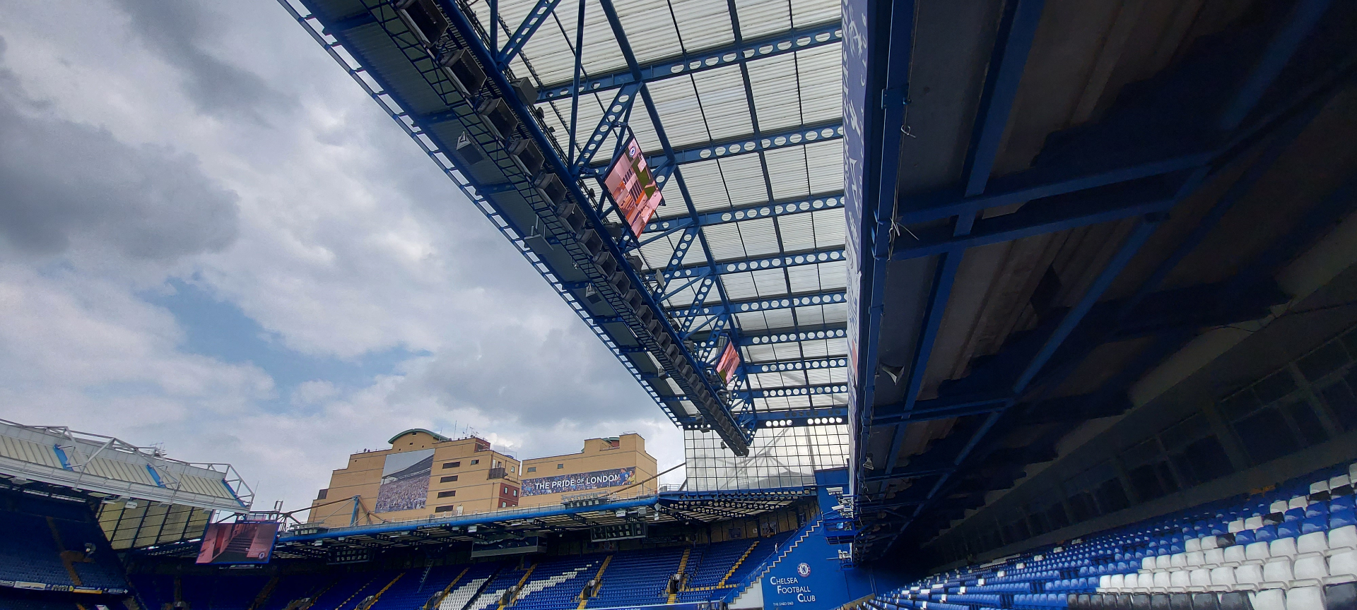 Chelsea FC — upward view from pitchside at the Shed End showing roof truss structure with suspended LED screen rigged at high level