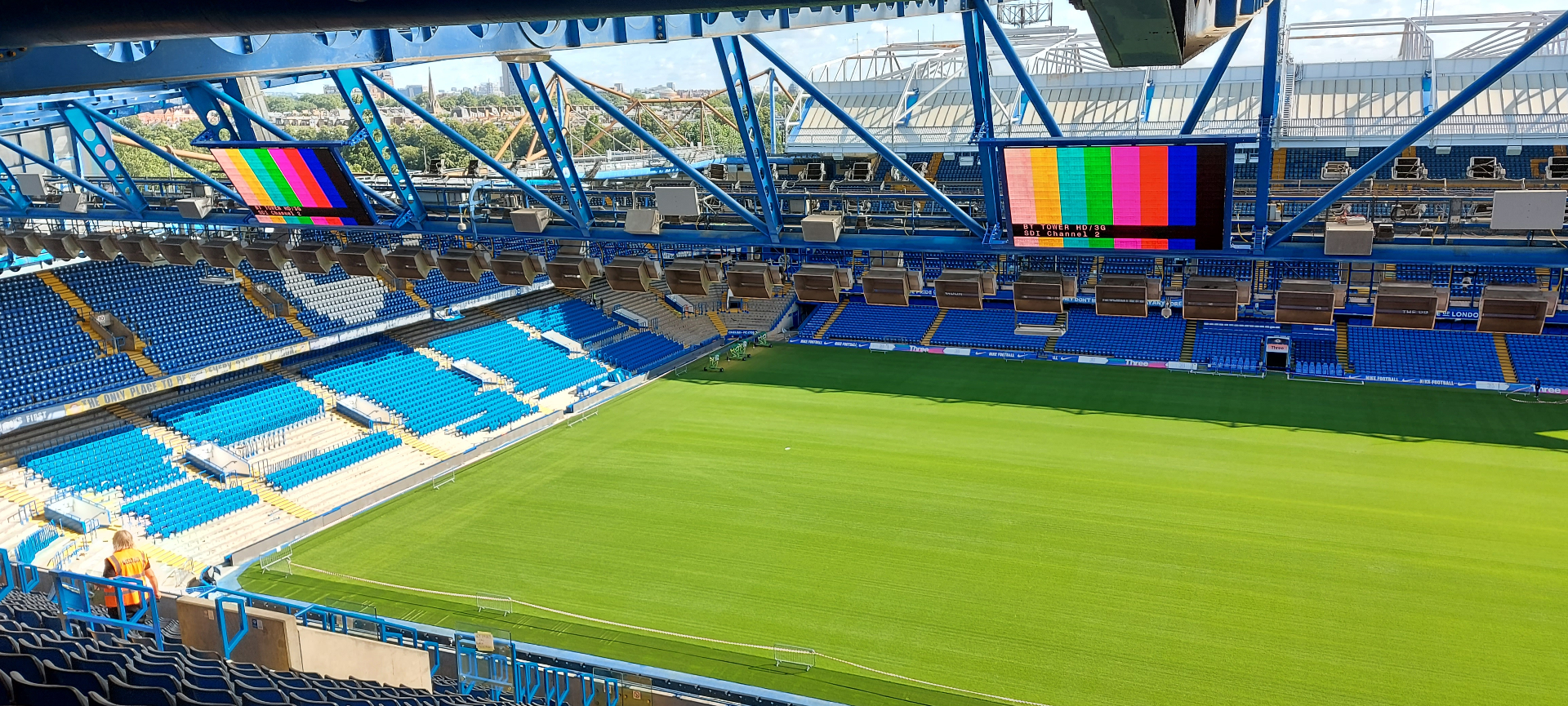 Chelsea FC — twin large-format LED screens displaying SMPTE colour bar test pattern during commissioning, viewed across empty Stamford Bridge bowl
