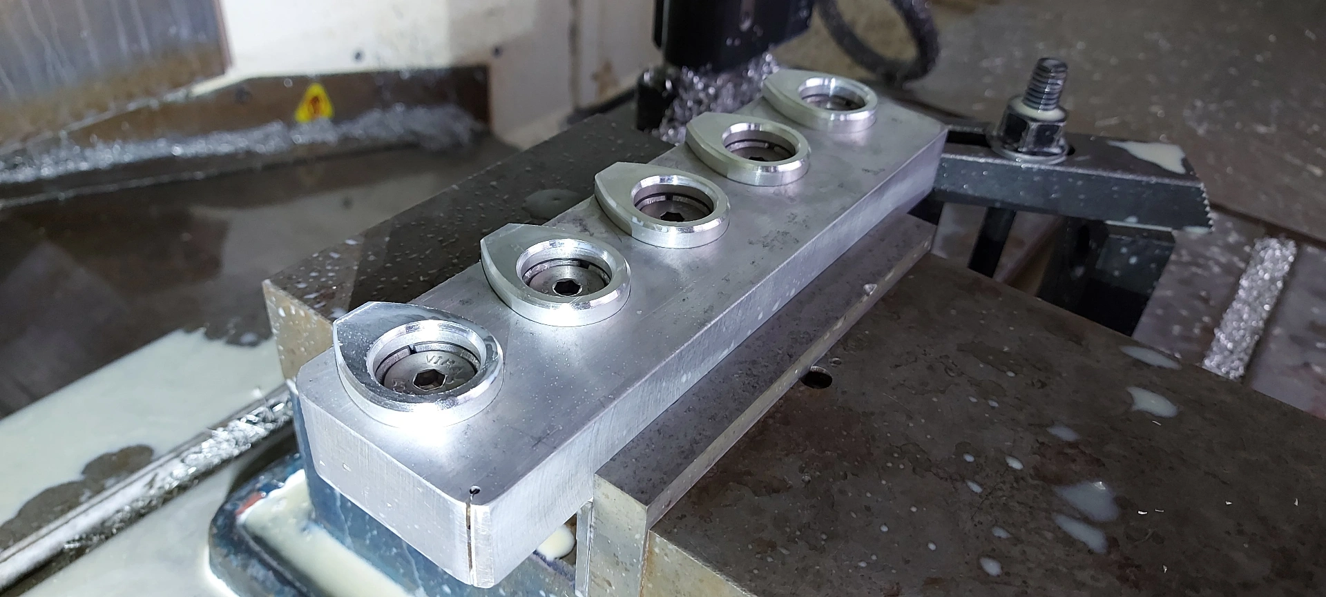 CNC — five-up machined component fixtured on CNC bed mid-cycle, showing concentric bored bosses and parallel toolpath finish