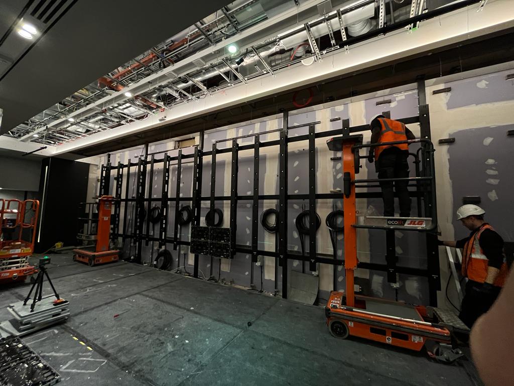 Schroders — bespoke steel sub-frame mid-install with engineers on cherry pickers, cable drops staged
